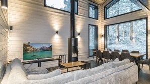 Een moderne woonkamer met een grote grijze hoekbank, een flatscreen televisie, een open haard en een eetgedeelte met een tafel en stoelen.