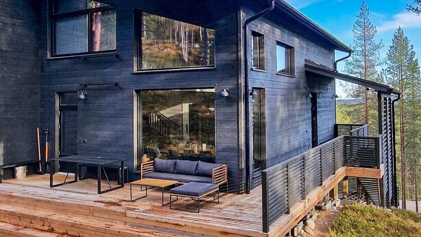 Een modern huis met twee verdiepingen, een donkere gevel, grote ramen en een houten terras met zitplaatsen.