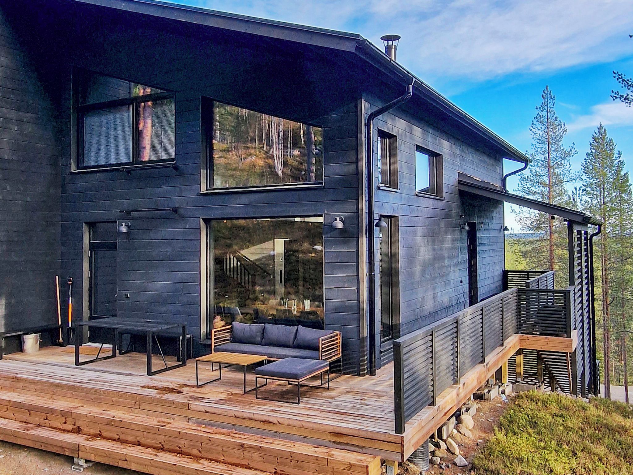 A modern two-story house with a deck, large windows, and a view of the surrounding forest.