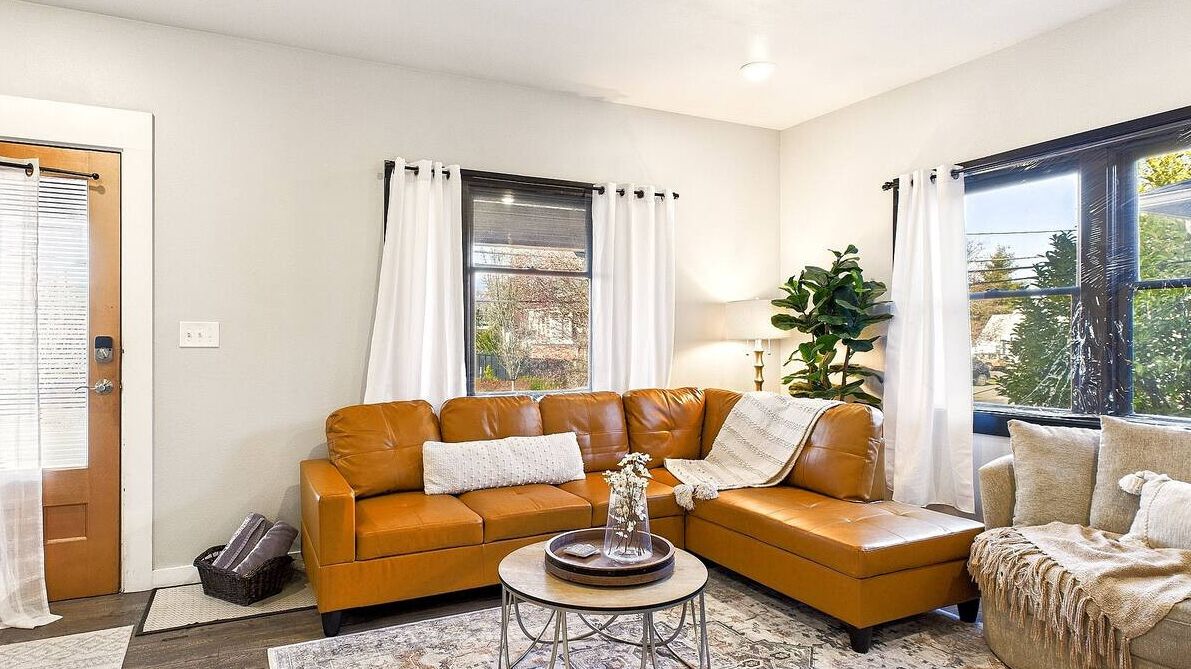 Un salon avec un canapé en cuir marron, une table basse et une grande fenêtre avec des rideaux.