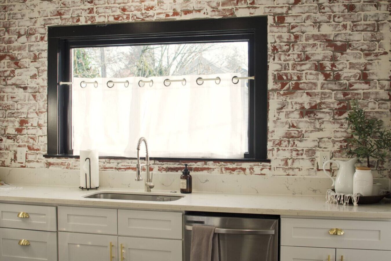 A kitchen with a brick wall, a window with a curtain, a sink, and cabinets.