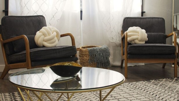 Due poltrone grigio scuro con struttura in legno e cuscini bianchi, simili a nodi, un tavolino da caffè rotondo con gambe dorate e un tappeto decorato su pavimento in legno.