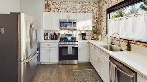 Una cucina compatta con mobili bianchi, elettrodomestici in acciaio inossidabile e una finestra che fa entrare luce naturale.