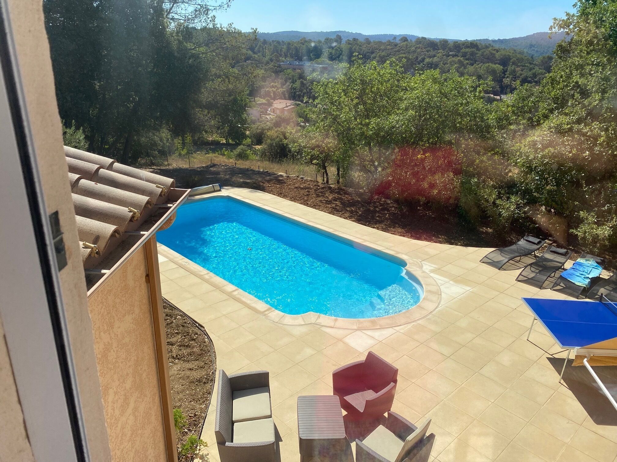 Une piscine rectangulaire, bordée d’une terrasse carrelée, entourée de chaises longues et d’une table, le tout se fondant dans un décor de verdure composé d’arbres et de collines.