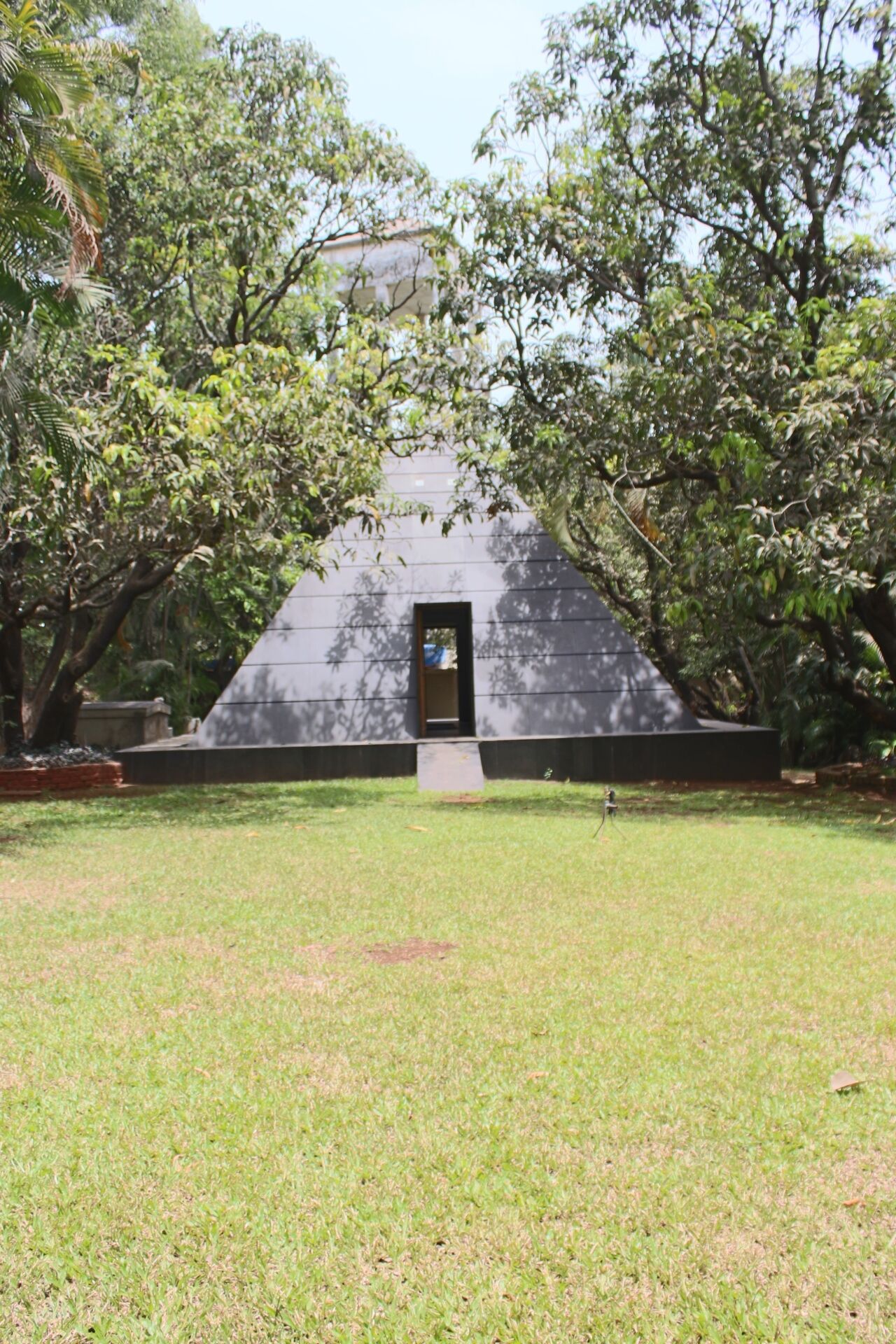 A grassy area with a triangular structure featuring a door, surrounded by trees.