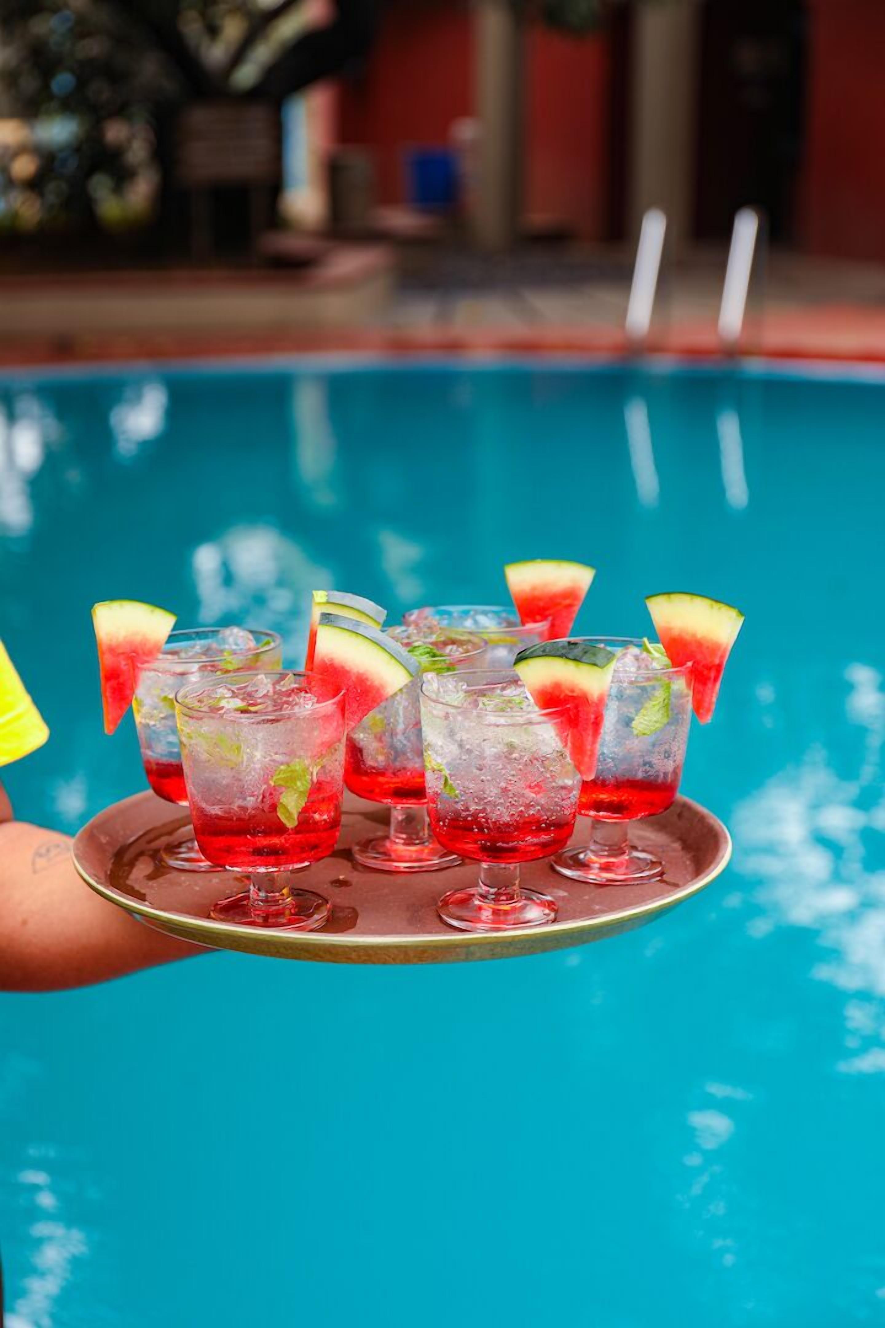 A tray with several glasses of red cocktails garnished with fruit slices, held over a pool.