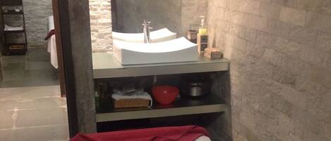 A massage room with a bed, a red towel, a white towel, a black cushion, and a sink with toiletries.