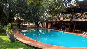 A swimming pool surrounded by lush greenery, with a wooden deck and a covered seating area. - Outpost (Alibag)