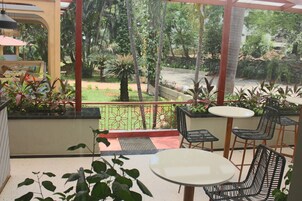 A conservatory with a glass ceiling, offering a view of a garden with various plants and trees. Inside, there are tables and chairs arranged for dining or relaxation.