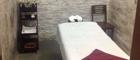 A small, narrow room with a bed, a chair, and a shelf with towels and a small plant.