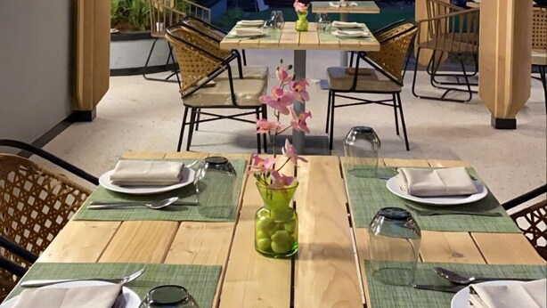An outdoor dining area with wooden tables and chairs, a central flower arrangement, and hanging wicker lamps. - Outpost (Alibag)
