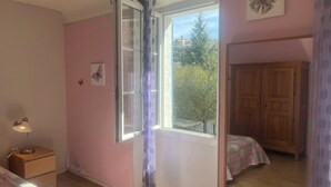 A room with a bed, a nightstand with a lamp, a wardrobe, and a window with a view of buildings and trees. - Views of Sault, cycling, hiking, Mont Ventoux (Sault)