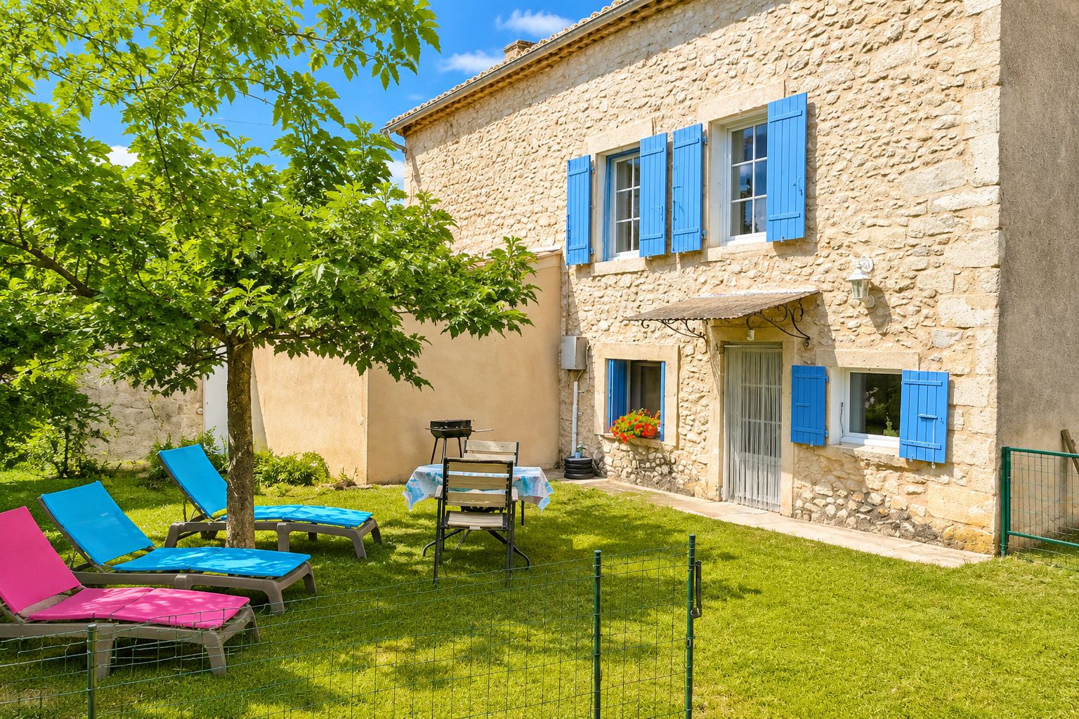 Un bâtiment en pierre avec des volets bleus, un espace extérieur aménagé avec des chaises et entouré d’une clôture, et un arbre.