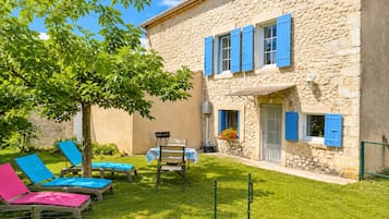 Un bâtiment en pierre avec des volets bleus, un espace extérieur aménagé avec des chaises et entouré d’une clôture, et un arbre.