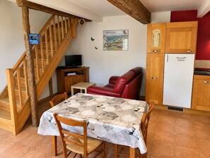 En matplats med ett bord täckt av en mönstrad duk, trästolar, en röd lädersoffa, en trätrappa, ett litet skåp med en tv och en tavla som hänger på väggen.