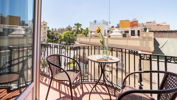 Un balcone con sedie in vimini e un tavolino, con vista su un panorama urbano che include edifici e palme.
