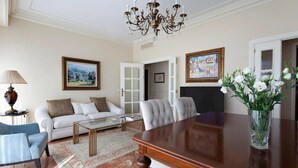 Un ampio soggiorno con un divano bianco, una poltrona blu, un tavolino in legno e un tavolo da pranzo con sedie. Si nota un lampadario, un quadro appeso alla parete e un vaso con fiori sul tavolo da pranzo.