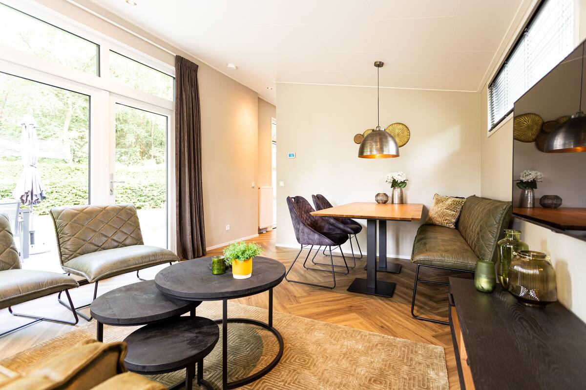 Ett modernt vardagsrum med en soffa, fåtöljer, ett matbord och ett soffbord. Det finns stora fönster med gardiner, en hängande lampa och ett trägolv.