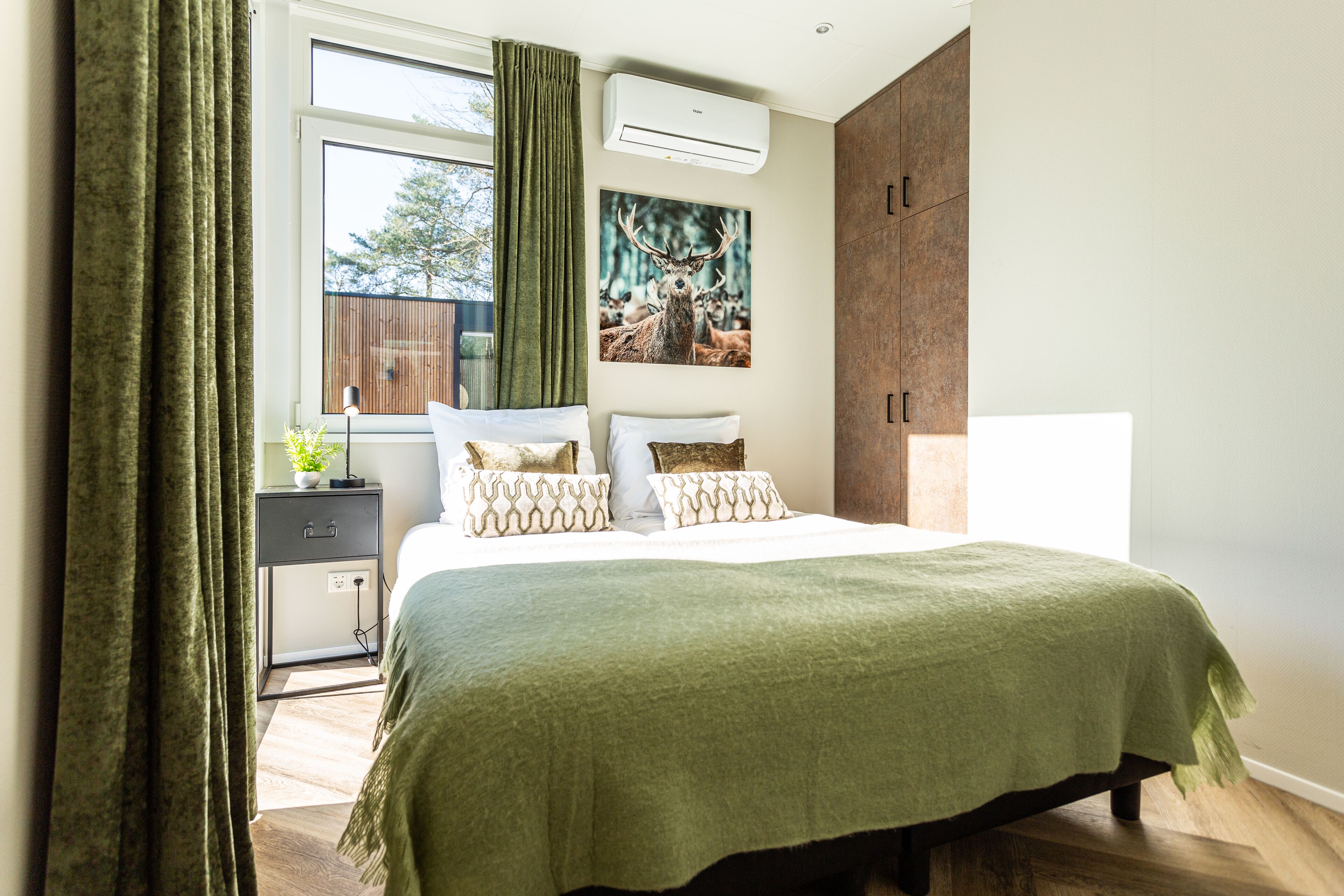 A bedroom with a bed, a nightstand, a window with curtains, a wall-mounted air conditioner, and a large wooden wardrobe.
