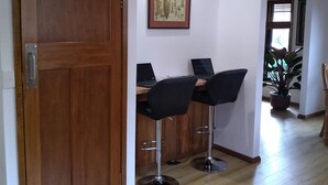 Una habitación con una puerta de madera, dos taburetes altos, un escritorio con dos computadoras portátiles, un cuadro en la pared y una planta en maceta en la esquina.