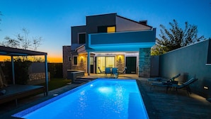 Una casa moderna de dos pisos con piscina, mobiliario exterior y una entrada bien iluminada.
