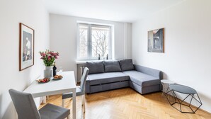 Een moderne woonkamer met een grijze hoekbank, een eettafel met stoelen, een klein rond tafeltje en een raam met jaloezieën.