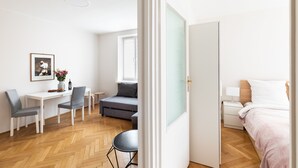 Een moderne, goed verlichte kamer met een houten visgraatvloer, een grijze bank, een klein rond tafeltje, een wit bureau met twee stoelen, een bed met wit beddengoed en een lijst met een afbeelding aan de muur.