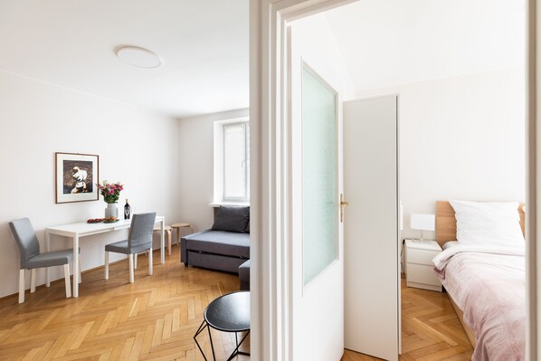 A modern, well-lit room with a wooden herringbone floor, a grey sofa, a small round table, a white desk with two chairs, a bed with white bedding, and a framed picture on the wall.
