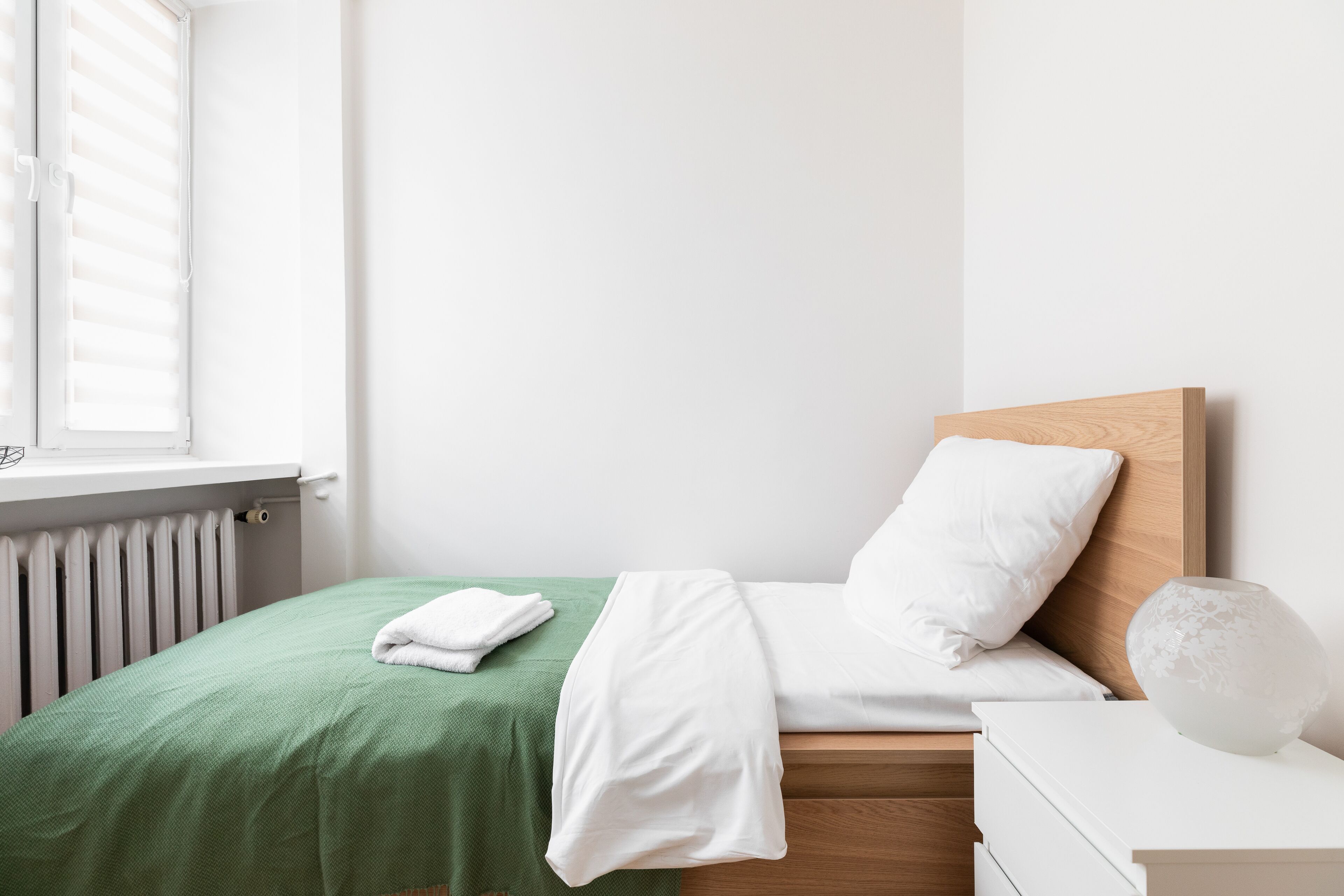A neatly made bed with a green bedspread and white linens, a wooden headboard, and a white bedside table with a decorative vase.