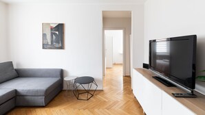 Een moderne woonkamer met een grijze bank, een klein rond tafeltje, een flatscreen televisie op een houten dressoir en een schilderij aan de muur.