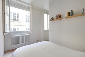 Une chambre à coucher avec un grand lit, une fenêtre dont le volet est ouvert, et deux étagères en bois remplies de livres.