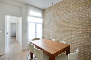 Un coin repas avec une table en bois et des chaises blanches, un mur en briques et une vue sur un couloir.