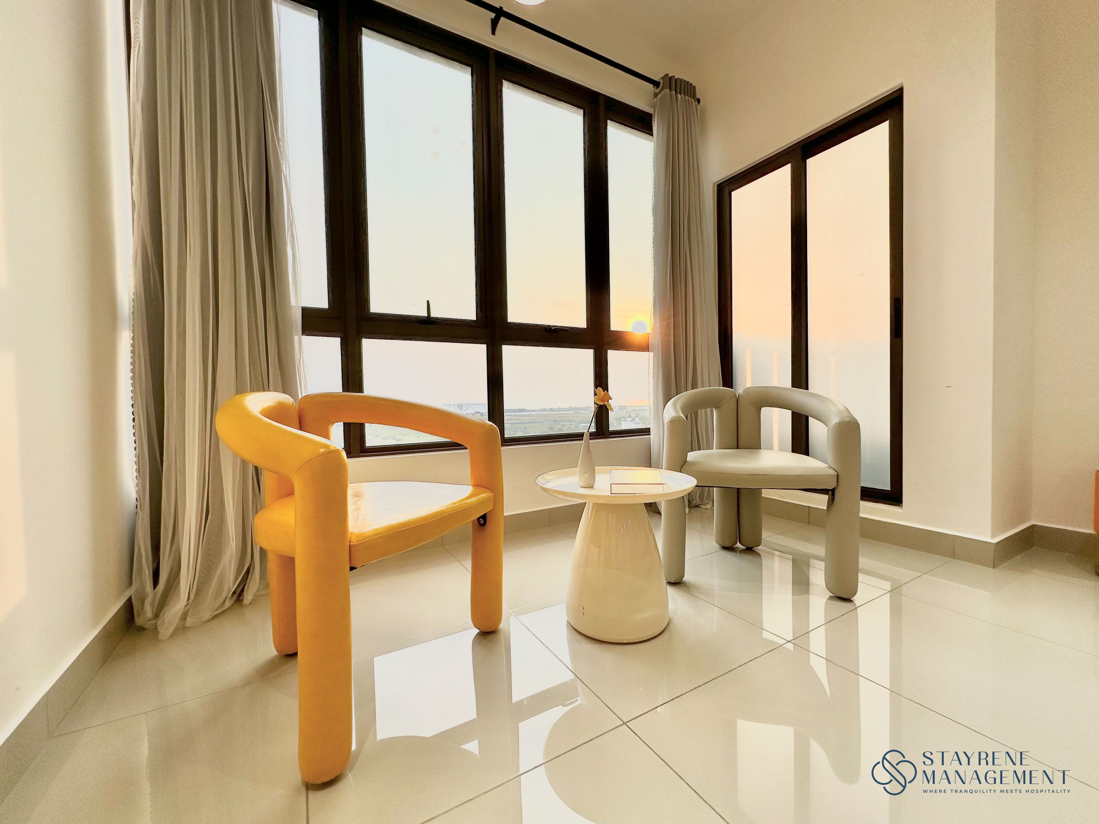 Una habitación de hotel moderna con ventanas amplias, dos sillones, una mesita y un florero con flores.
