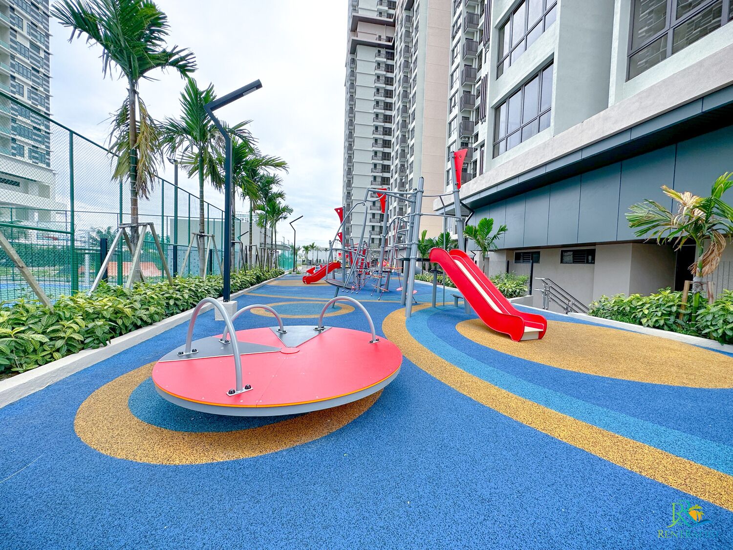 A playground with slides and climbing structures, surrounded by a fence and palm trees, in an urban residential area.