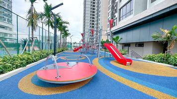 A playground with slides and climbing structures, surrounded by a fence and palm trees, in an urban residential area.