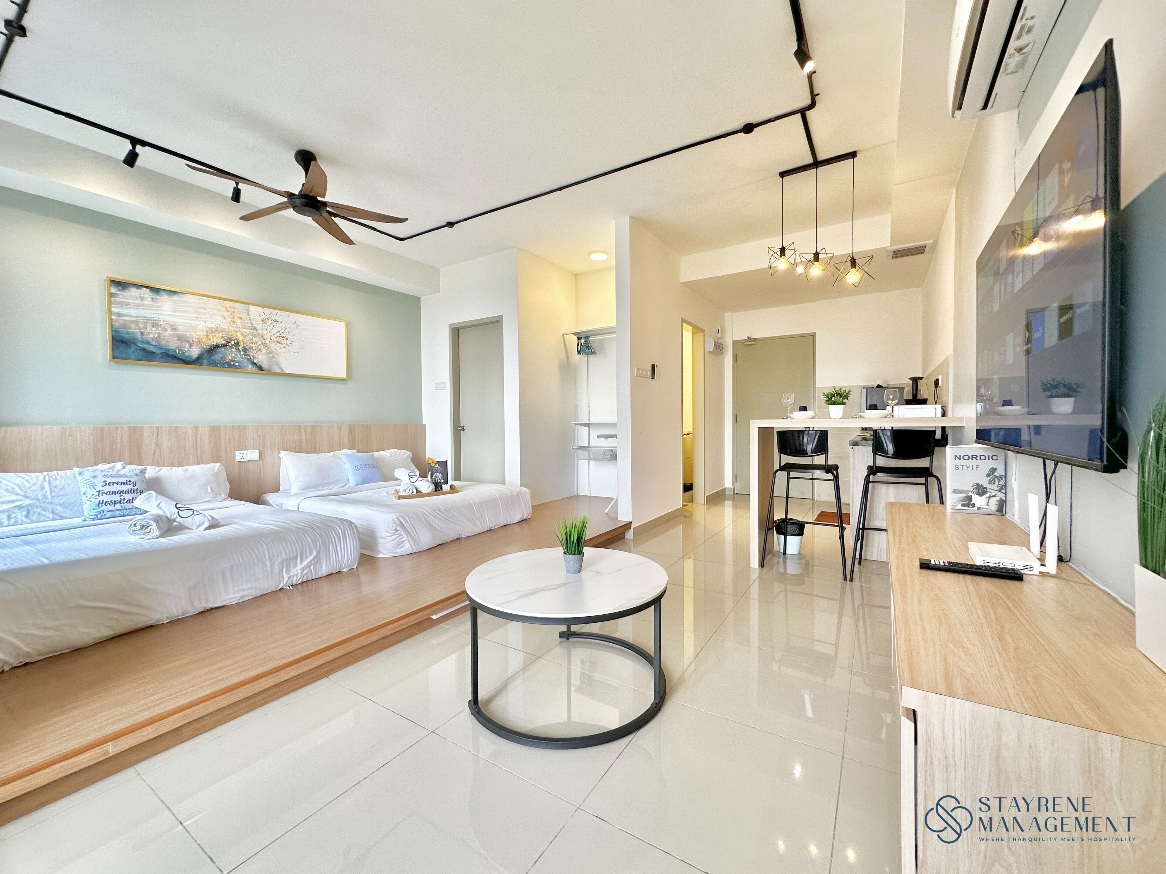 A modern hotel room with a bed, a sofa, a small table, and a TV. There is a kitchen area with a counter and chairs.