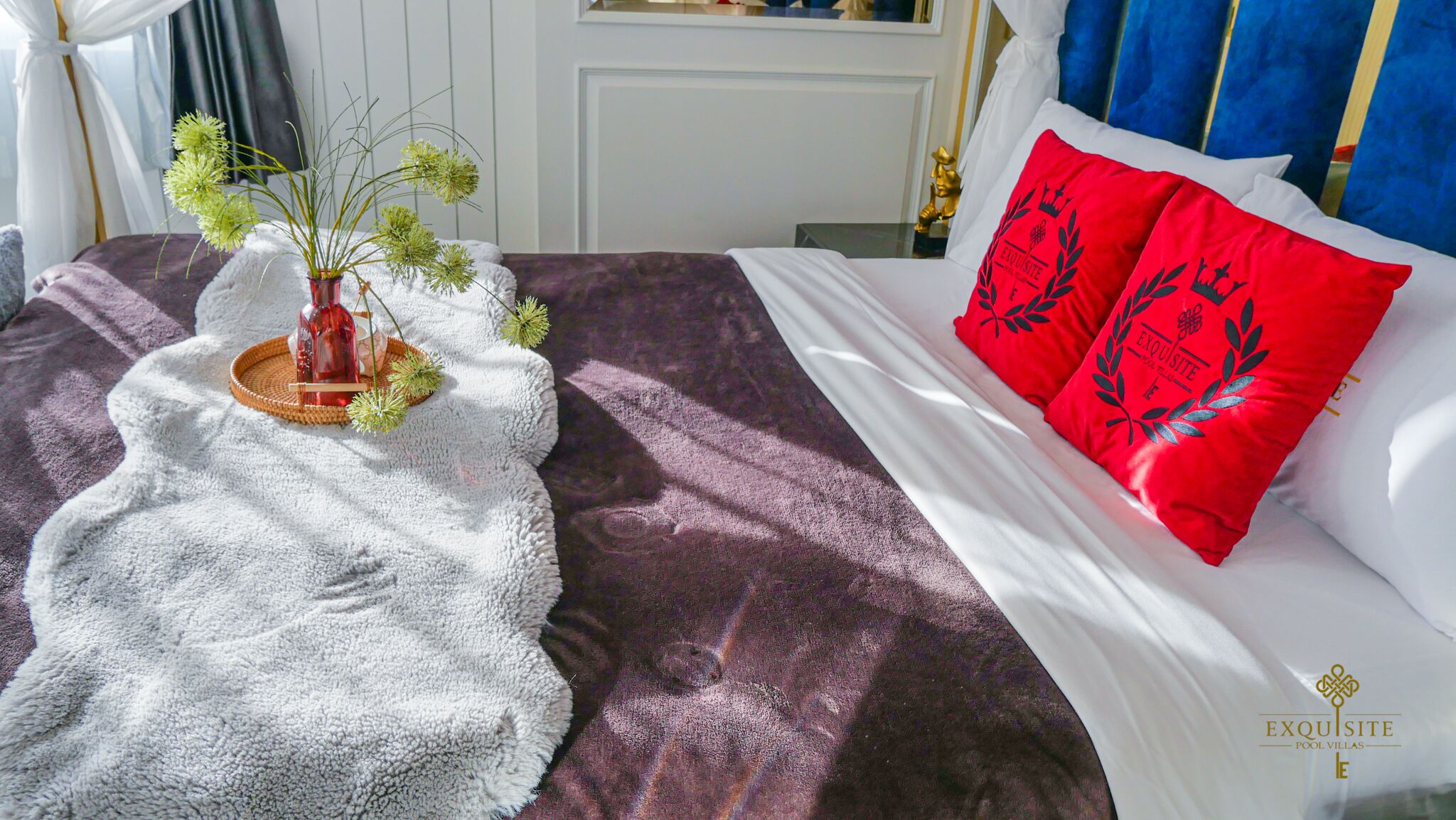 Una cama bien hecha con una colcha blanca y un almohadón rojo con un diseño de laurel.