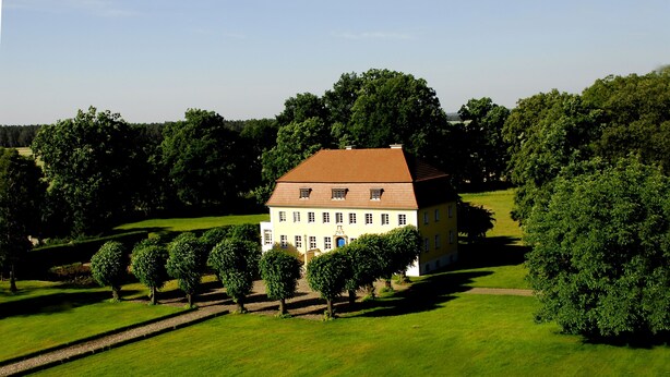 Ein großes, einstöckiges Gebäude mit symmetrischer Fassade, umgeben von einer gepflegten Rasenfläche und einigen Bäumen.