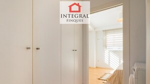 Una habitación moderna y minimalista con paredes blancas y suelo de madera. Cuenta con un armario, una cama con colcha a rayas y un electrodoméstico empotrado.