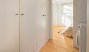 Quarto moderno e minimalista com paredes brancas e chão de madeira. Possui um guarda-roupa, uma cama com colcha riscada e um eletrodoméstico embutido.