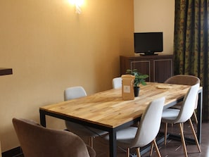 Un coin repas avec une table en bois, quatre chaises, une plante en pot, un téléviseur posé sur un meuble et un mur agrémenté d’un luminaire.