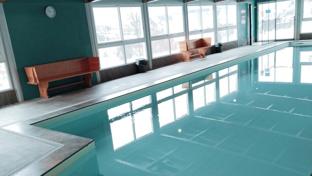 An indoor swimming pool with clear water, a wooden bench along the edge, and large windows providing natural light. - Duplex T3 in Jausiers - 6 people, terrace, parking (Jausiers)