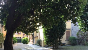 A courtyard with a pathway, trees, and buildings with shutters. - Apartment 'In Villa Belli' with Shared Garden & Wi-Fi (Miasino)