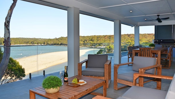Un balcon spacieux offrant une vue sur une plage et une rivière. On y trouve des meubles en bois, notamment une table et des chaises, ainsi qu’une plante en pot.