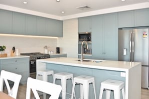 A modern kitchen with light blue cabinets, a stainless steel refrigerator, a built-in oven, and a central island with bar stools. - BEACH AT THE BOTTOM OF YOUR GARDEN & OCEAN VIEWS PACIFIC CRES (Sutherland Shire Council)