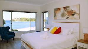 Une chambre avec une grande fenêtre, un lit recouvert de draps blancs, un fauteuil bleu et un tableau accroché au mur.