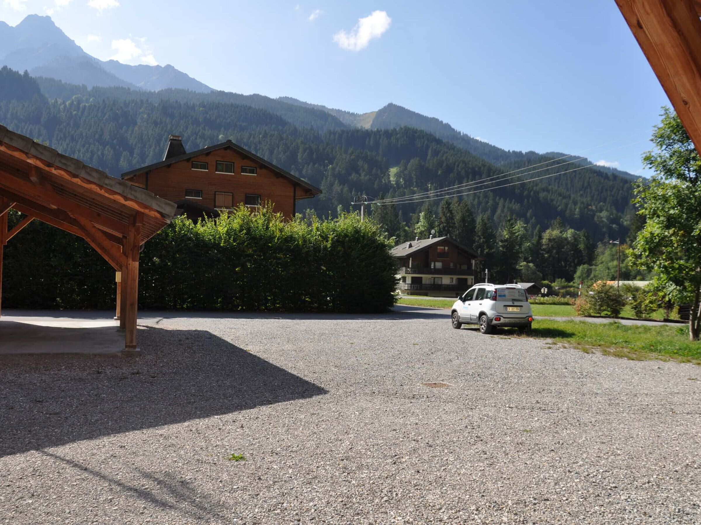 A building with a wooden structure in the foreground, a gravel driveway, and a white car parked in the distance.