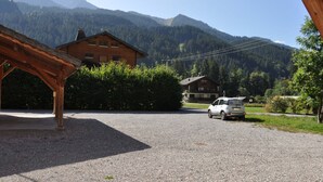 A building with a wooden structure in the foreground, a gravel driveway, and a white car parked in the distance. - Charming apartment with terrace, covered parking and WIFI (La Chapelle-d'Abondance)