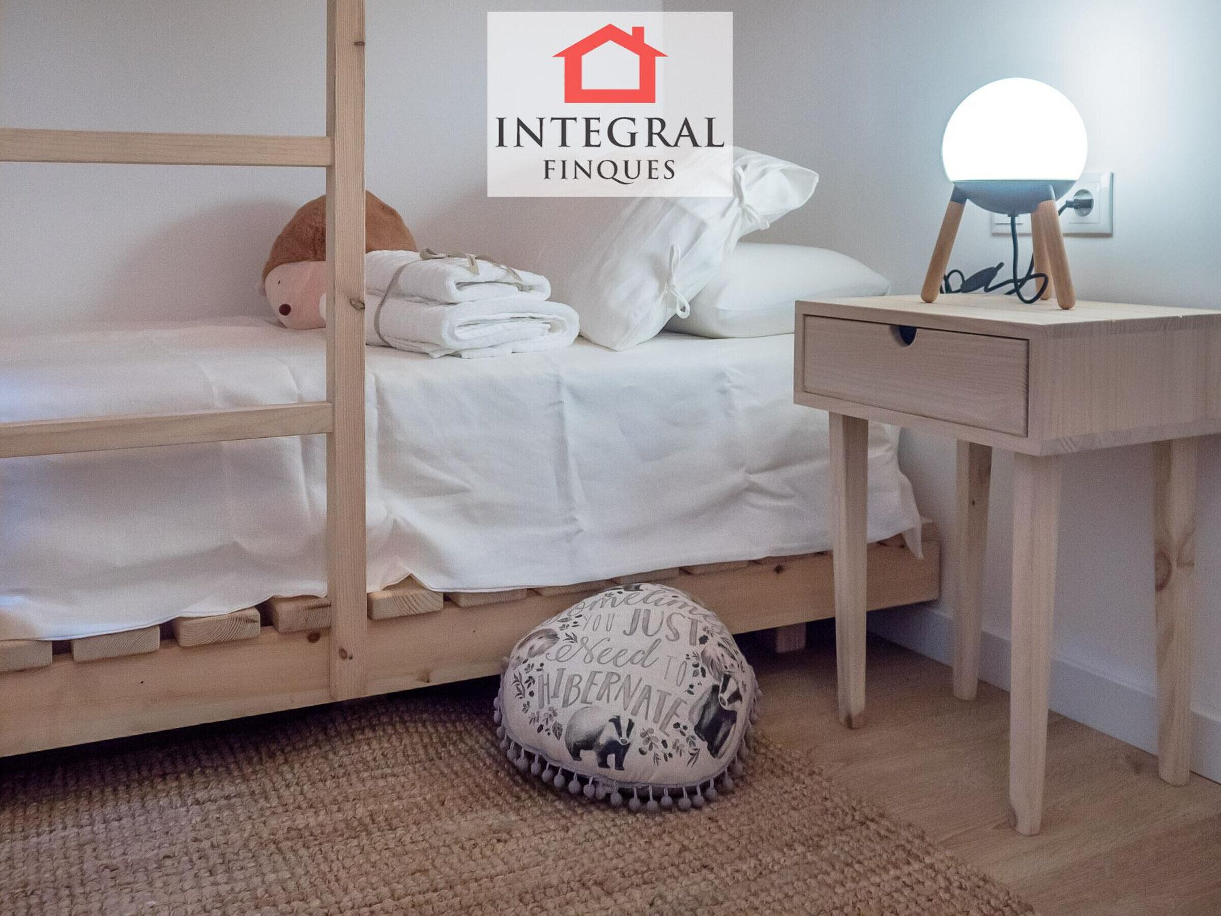A bedroom with a bunk bed, a bedside table with a lamp, a small ottoman, and a wall-mounted light.
