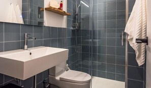 A modern bathroom with a sink, toilet, and shower. There is a towel rack and a shelf with toiletries. The walls are tiled in a dark color. - Modern duplex in Palamós center with terrace and private garage (Palamós)